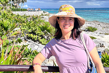 Kun Yang smiling on a coastal boardwalk, wearing a sun hat and lavender dress, with ocean and tropical greenery in the background.