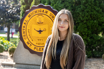 Kylie Pearce has waist-length light brown hair with blonde highlights. She is wearing a taupe cardigan over a black shirt and stands in front of the CMU seal in the Warriner Mall.