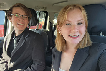Colten Lambart, left, and Cailin Lambart, right, pose for a selfie together in a car with a dark interior. Colten has a coordinating dark gray jacket, shirt and tie. Cailin wears a dark gray jacket over a white shirt.