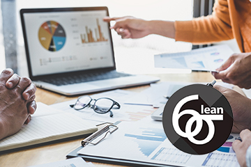 A laptop displays pie charts and bar graphs and reports with similar graphs are all on a table with people gesturing to the data. In the corner is the Lean Six Sigma logo.