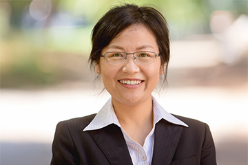 Li Zhang stands outdoors and faces the camera and smiles. She has black hair that is pulled back. She wears wire frame glasses. She has on a black blazer and a white button down shirt with dark pinstripes.