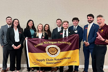 A group of nine students and their faculty advisor - who is showing a peace sign - stand in a hallway with a maroon and gold CMU Supply Chain Management Association flag. They are wearing business professional clothing.