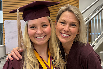 A student with long blond hair poses in a cap and gown with Lori Driessnack who has shoulder length blond hair and wears a maroon shirt.