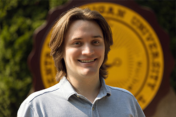 Lucas Sherman has shoulder length hair and wears a gray polo shirt and stands in front of the CMU seal on the Warriner Mall.