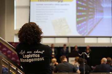 A woman with dark curly hair stands with her back to the camera. She has a black shirt with white writing that reads "Ask me about Logistics Management" and she's watching four students give a presentation in the Grawn Atrium.