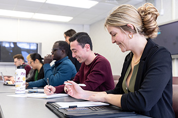 Students sit in a classroom during a lecture. In the foreground a young woman with blond hair in a bun laughs and looks at her padfolio. A faculty member can be seen chatting with students in the background.