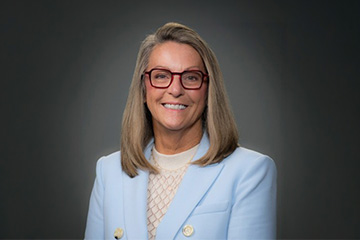 Melanie Kalmar smiles in a professional headshot wearing a light blue blazer and red glasses against a dark background.