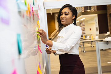 A student in a maroon pencil skirt and white button down shirt places colorful post-it notes on a white board in the Grawn atrium.