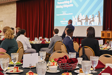 Students dressed in business professional clothing sit at tables in the UC rotunda with salads, drinks and desserts in front of them. A speaker stands at the front of the room in front of an out of focus display screen.