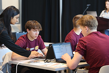 Two highschool students in maroon t-shirts face their laptops as a mentor with long dark hair and a black shirt leans over to offer advice.