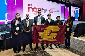 Six CMU logistics students stand with a maroon Action C flag at the National Grocers Association conference where they competed and placed second in a logistics competition.