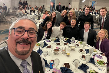 Nick Hussein takes a selfie at a PSE event. He has a neat goatee, salt and pepper hair and black glasses. He is wearing his signature vest over a collared shirt and tie. Behind him is a large group of smiling students at a table.