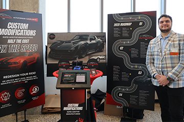 A bearded student in a plaid blazer and black pants stands in front of a multimedia display showing custom car modifications created by his venture.