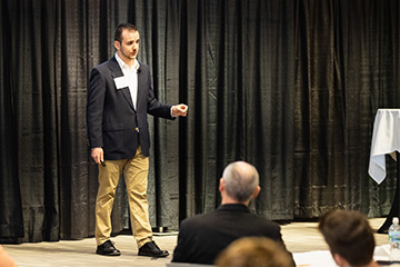 A student in a black jacket, white shirt and khaki pants presents in front of a black curtain. He has a remote in his hand to advance his slides. People sit in the foreground and watch him.