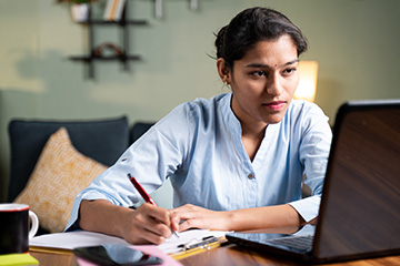 A woman in a long-sleeve blue shirt holds a red pen and takes notes while looking at a laptop.