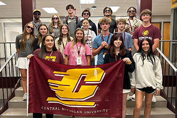 Incoming freshman students stand with their peer advisors near the Business Student Services office. They are dressed in summer clothes, mostly shorts, tshirts and sweatshirts. Some wear sunglasses. They hold an Action C flag in maroon in gold in front.