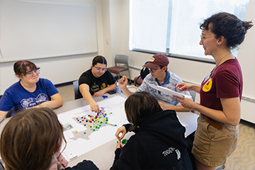 A groups of students sit around a table in the Park Library. On the table is a map of the United States with colorful red, blue, yellow and green game pieces. Another woman stands overlooking the table holding papers. She wears a CMICH Press tshirt.