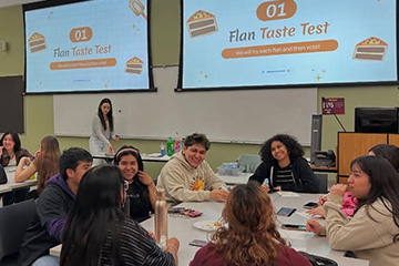 Students sit at a table in a classroom with paper plates and drinks in front of them and various handouts. They are smiling, talking and eating. The projector screen in the background above them reads "Flan Taste Test."