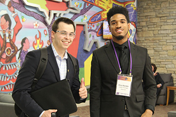 A student in a black blazer holding a black portfolio gives a thumbs up sign and stands next to a student in a black blazer with a nametag in front of a wall mural.