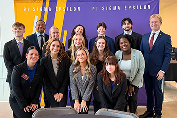 Fourteen students, a mix of men and women, stand in front of a purple and yellow Pi Sigma Epsilon backdrop. They are all wearing business professional clothing and posing as a group.