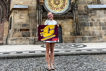Reghan Haley holds a maroon and gold CMU flag beneath Prague’s ornate astronomical clock.