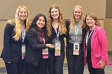 Four female students stand together in a line. Three have long blond hair and one has long dark brown hair. They are all wearing black business suits and lanyards. One holds a crystal trophy. The advisor is at the right side in a bright pink jacket.
