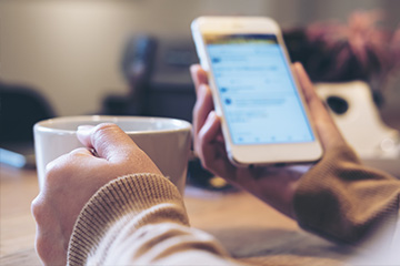 A woman holds a ceramic coffee cup in her left hand and a phone in her right hand that appears to display a social media feed.