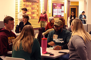 Students gather in Grawn Atrium to meet their teams and practice their pitches for the Spark Challenge. In the foreground is a table with four students discussing their Impact Path prompt. Other tables have groups of students in conversation.