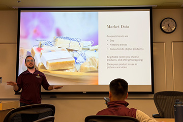 Daniel Lahey stands in front of students in the 150G classroom. He is wearing a maroon poly shirt. Students sit at tables throughout the room and face him. Behind him is a pull-down screen with a presentation slide projected on it.