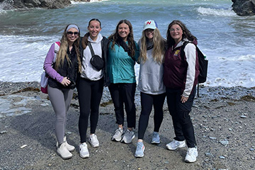 Five students stand on a beach in the United Kingdom. They are wearing jackets, sweatshirts, leggings and tennis shoes. The beach is rocky and waves roll in behind them.