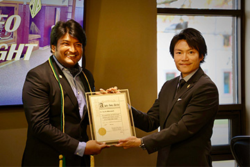 Surith Bhowmick receives an Alpha Iota Delta certificate from Edward Chou. Both are dressed in jackets and Bhowmick wears green and cold cords and tassels.