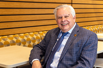 Thomas Anthony sits at a booth table in Grawn Hall. He's wearing a dark blue plaid jacket with a white shirt and blue tie. He has white hair and is smiling.