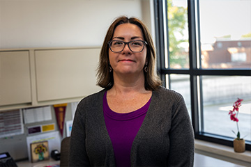 Tracey DeFeyter stands in her office and smiles at the camera with a closed smile. She wears a purple shirt, a gray cardigan and black framed glasses. Her hair is shoulder length and brown. She has dangly earrings.