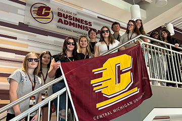 A large group of students pose on the stairway and balcony in the Grawn Hall atrium. They are wearing lanyards and many have on sunglasses. They are holding an Action C flag. On the wall behind them is a College of Business logo.