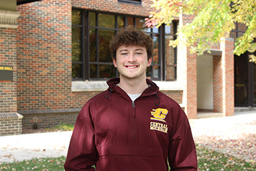 Tyler Harvill smiles outside Grawn Hall at Central Michigan University, wearing a maroon CMU jacket with the university's gold logo.