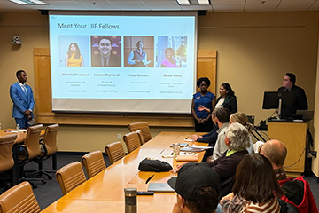 UIF students present to stakeholders in a Park Library conference room. The students stand at the front of the room near a display screen with their bios. Faculty and staff members sit at tables in front of them and listen intently.