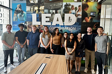 A group of students in the Wakeling-Gendron Entrepreneurial Scholars program pose with LEAD Venture Corporation representatives in a boardroom during a visit. In front of them is a large table and behind them is a wall graphic that reads "LEAD".