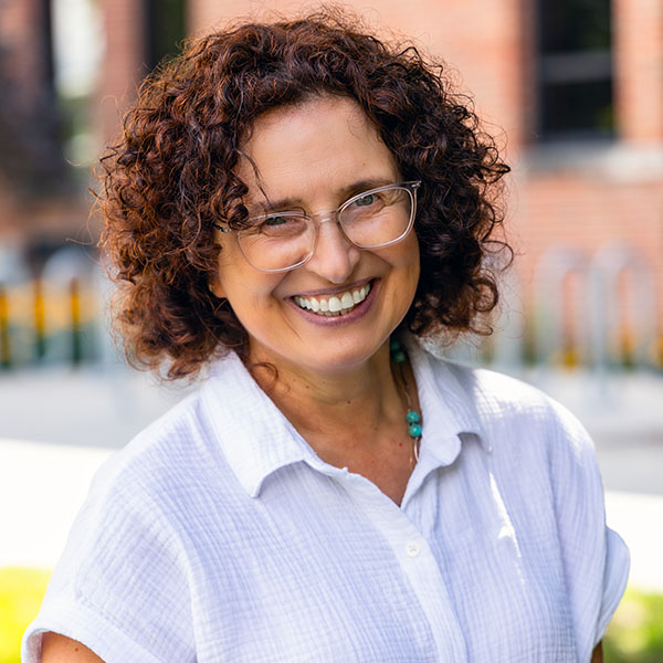 Crina Tarasi poses outside of Grawn Hall. She has shoulder length curly hair, clear framed glasses and a big smile toothy that creates smile lines around her eyes. She is wearing a white linen button down shirt and a silver and turquoise necklace.