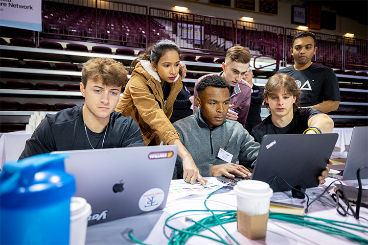Six students gather around laptops in Finch Fieldhouse. They are pointing and talking intently as they compete in the ERPsim competition. The table is covered in papers, network cables and coffee cups.