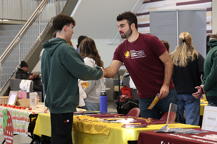 A student in a green hoodie and black pants shakes hands across a table with swag and brochures with a logistics department representative wearing a maroon and gold t-shirt at the CBA Spring Involvement Fair.