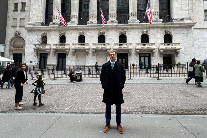 Adam Rose stands in front of the New York Stock Exchange in New York City, dressed in professional attire.