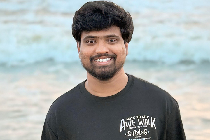Akhilreddy Bokka faces the camera and smiles. He is wearing a black t-shirt with non-sensical English text. The ocean appears behind him and is out of focus.