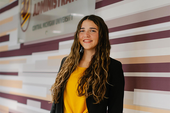 Ainsley Reigler wears a black blazer over a gold shirt and poses at the top of the stairs in the Grawn atrium.