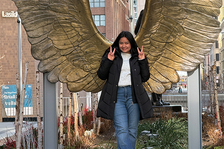 Akanksha Tyagi stands in front of large golden metal wings as she poses for a photo. She's wearing a black winter jacket, sweater and jeans. She's smiling and making peace signs with her hands.