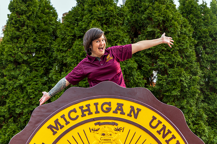 Alisha Draper sits on top of the CMU seal in Warriner Mall. Her hands are outstretched and she has an exaggerated grin. She wears a maroon CMU polo shirt. She has short brown hair.
