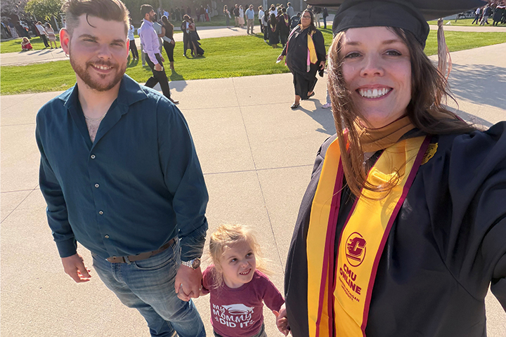 CMU MBA graduate Alyssa Welch smiles in her cap and gown while taking a selfie with her husband and young daughter after commencement.