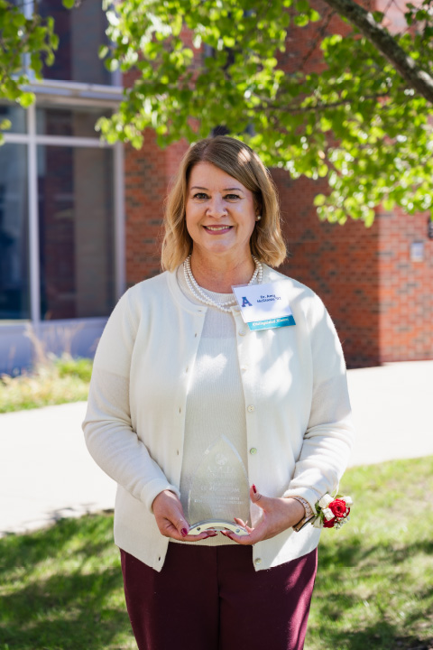 Amy McGinnis smiles outdoors while holding a clear award in front of a brick building.