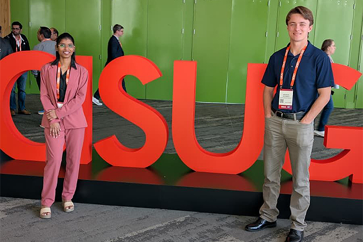 Jaahnavi Nagaraju wears a pink suit and black shirt, standing alongside Brenden Haldeman, who wears a blue shirt and khaki pants, at the ASUG Tech Connect event. They are standing in front of red 3D sculpted letters that spell out 