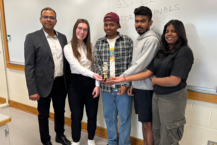 Associate Dean Sanjay Kumar stands with the I-Ride student team in a classroom as they hold an award together. The group is positioned in front of a whiteboard, representing their project work in BIS 636.