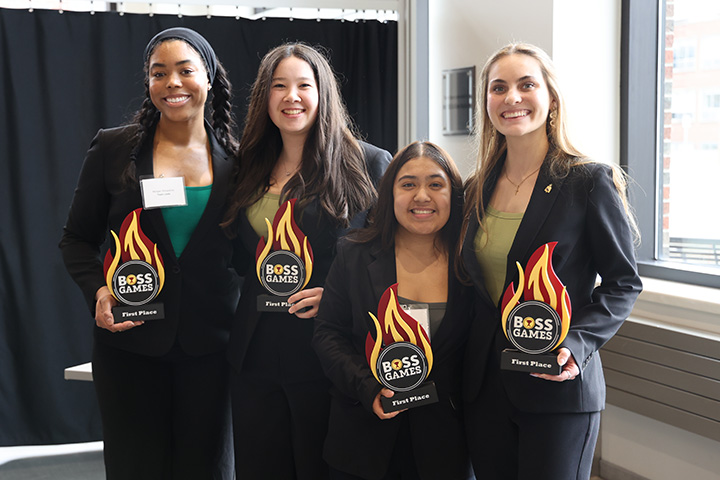 Four students hold first-place Boss Games awards and smile at the camera during a College of Business competition event.